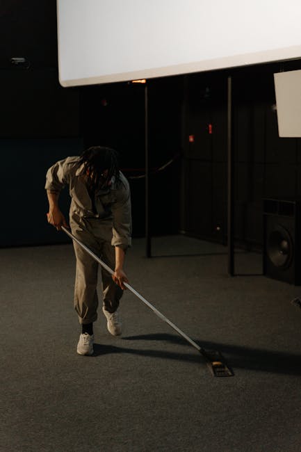 A close-up view of a professional vacuum cleaner with a red top and transparent dust container emitting bubbles, positioned on a pink carpeted floor in a room with white walls. The vacuum's black and 