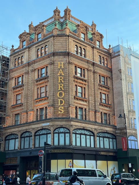 Photograph of the historic Harrods building in Knightsbridge, London, showcasing its ornate stone facade with detailed architectural features and large arched windows on the ground floor. The building
